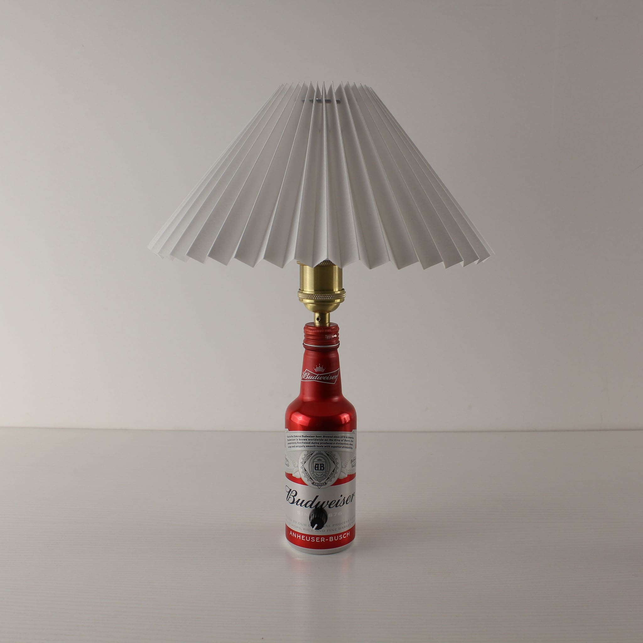 A cordless Budweiser aluminum bottle lamp with a white pleated lampshade, shown turned off in a clean studio setting