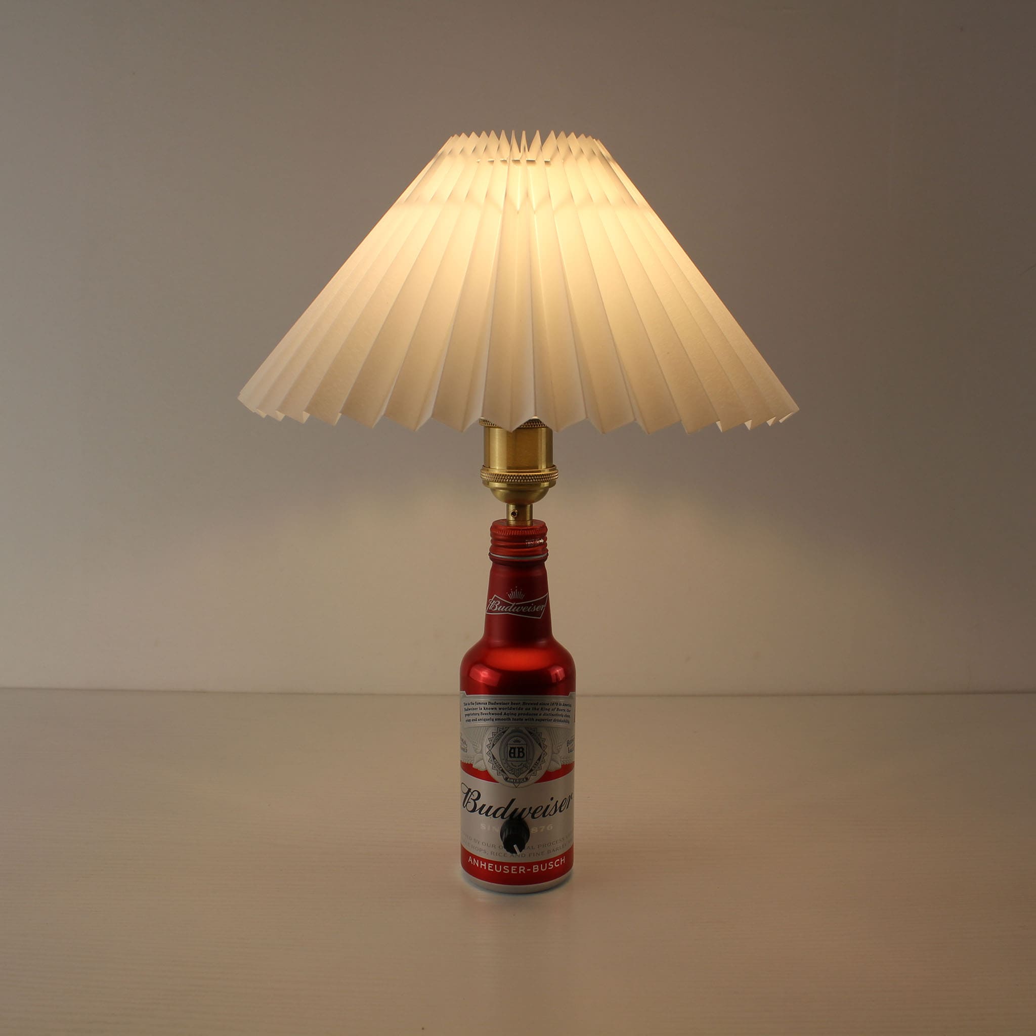 The Budweiser cordless lamp turned on, emitting a warm 2700K glow through a white pleated shade
