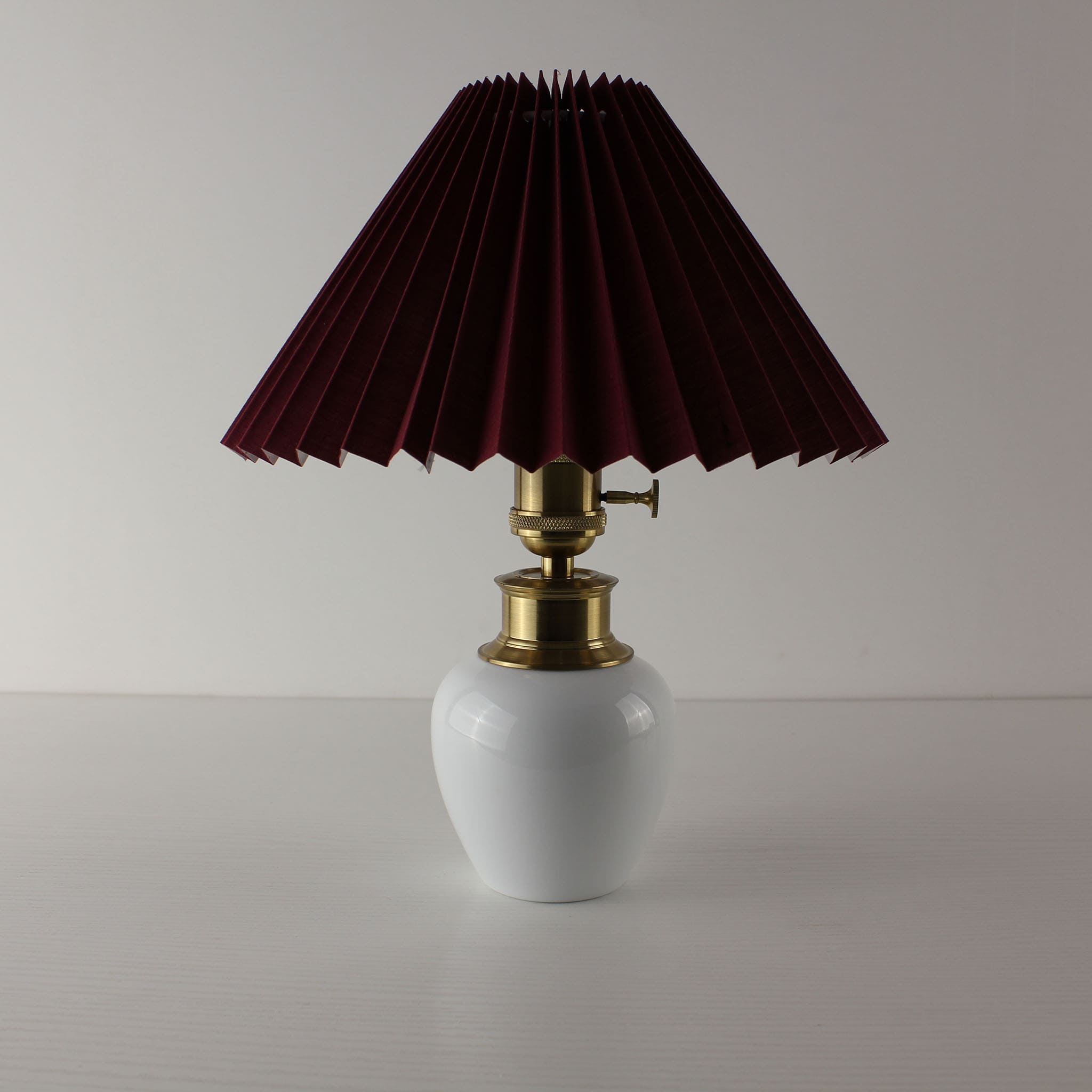 Vintage style portable table lamp featuring a white porcelain base and a burgundy pleated lamp shade.