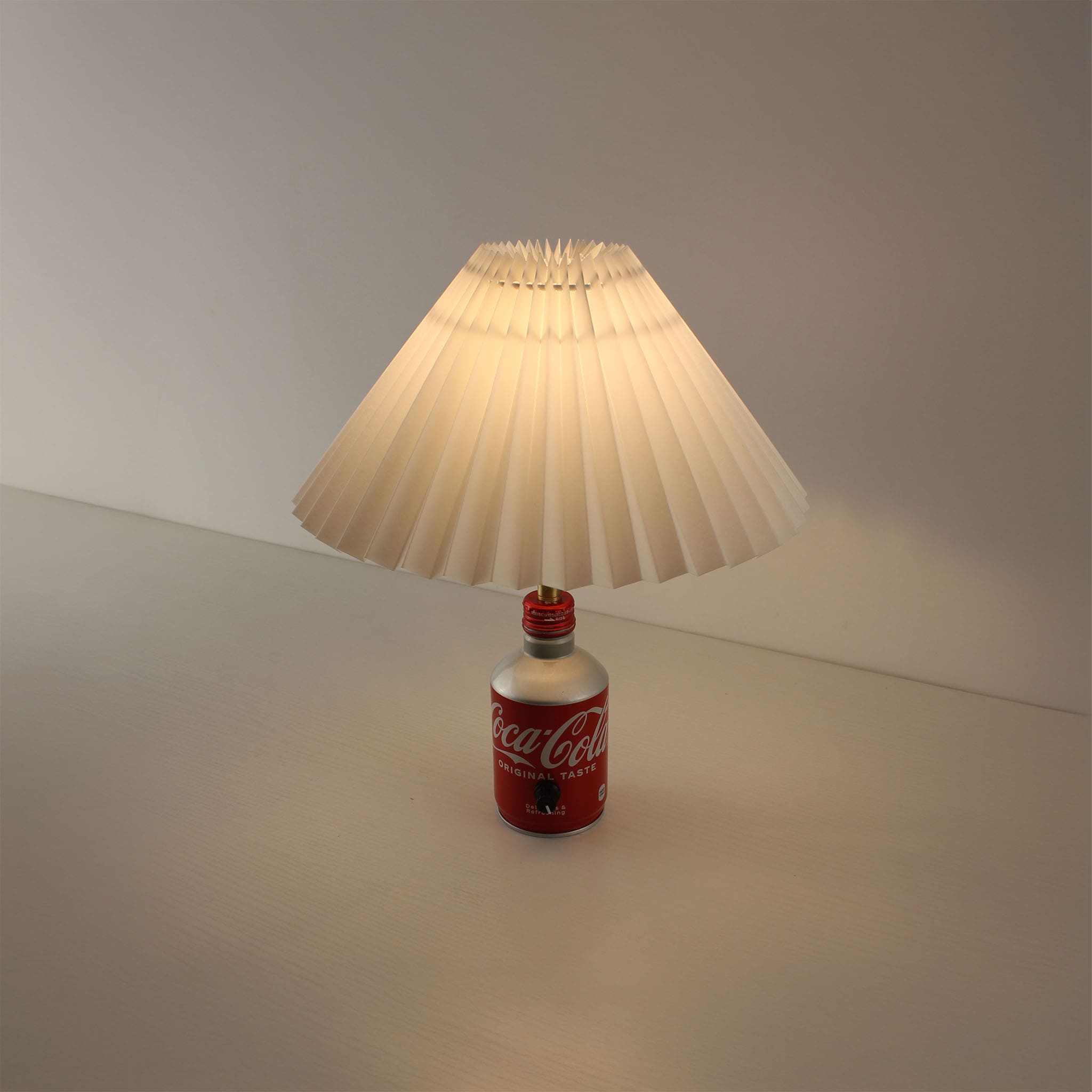 A Coca-Cola bottle table lamp turned on, emitting a warm, soft glow through its white pleated shade in a clean, contemporary setting.