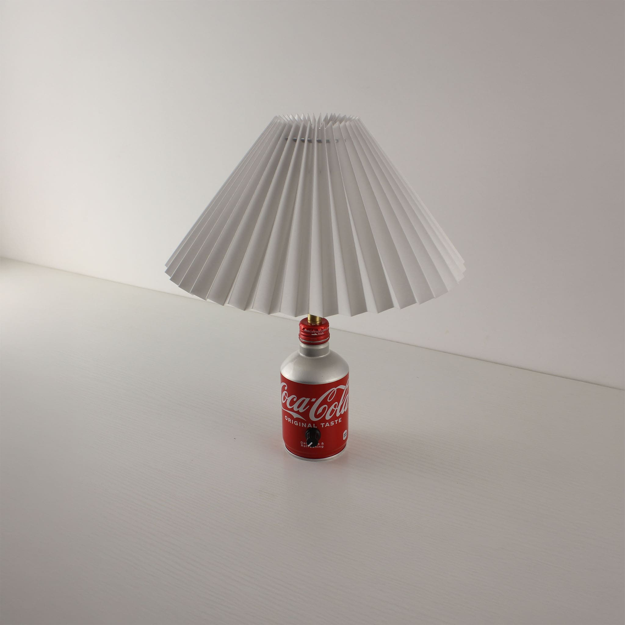 A desk lamp featuring a red and silver Coca-Cola bottle base with a white pleated lampshade, shown turned off against a minimalist white background.