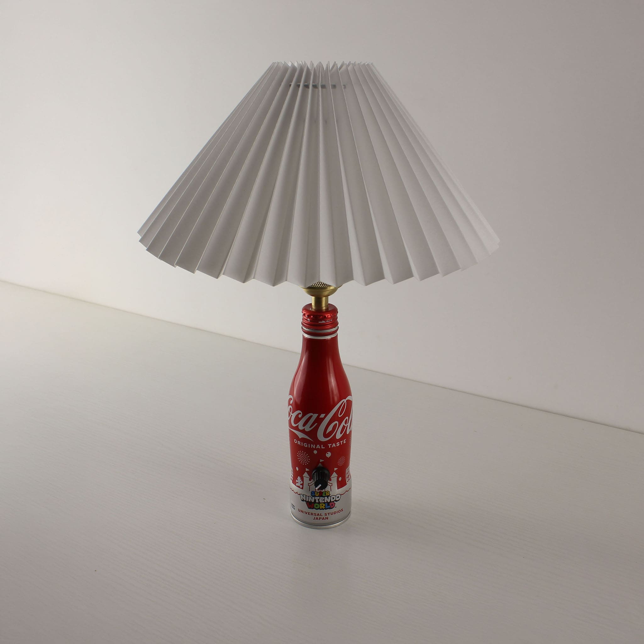 A compact version of the Coca-Cola bottle lamp using the "Original Taste" aluminum bottle design with a white pleated shade