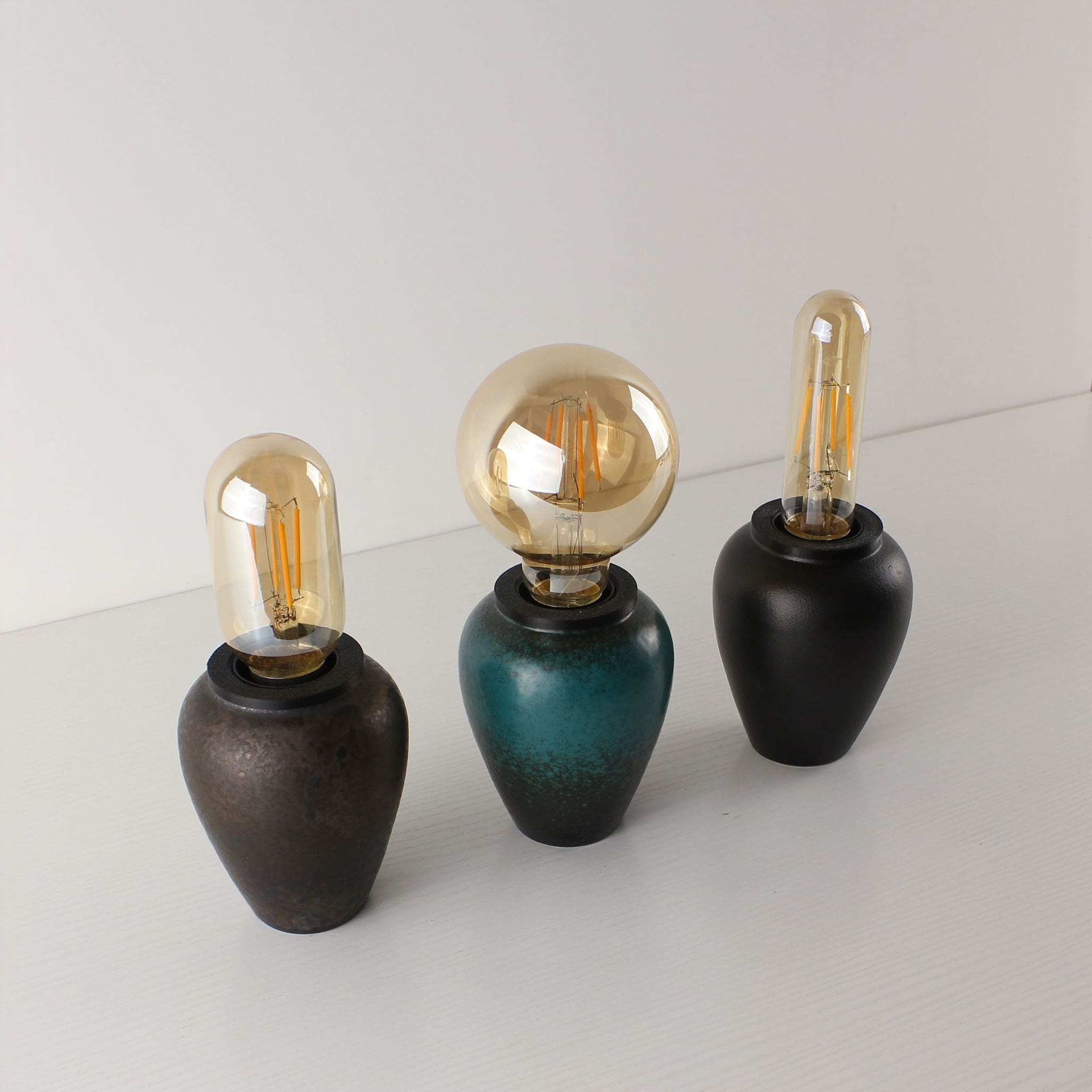 Side-by-side comparison of three cordless ceramic lamps in Industrial Rust, Kiln-turned Green, and Matte Black finishes, each paired with a unique Edison LED bulb