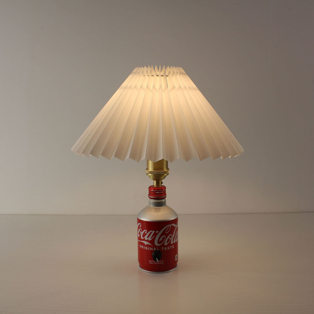 Front view of a cordless lamp made from a Japanese Coca-Cola bottle with a white pleated shade turned on, emitting warm light.