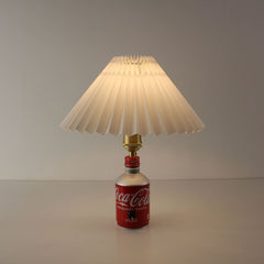 Front view of a cordless lamp made from a Japanese Coca-Cola bottle with a white pleated shade turned on, emitting warm light.