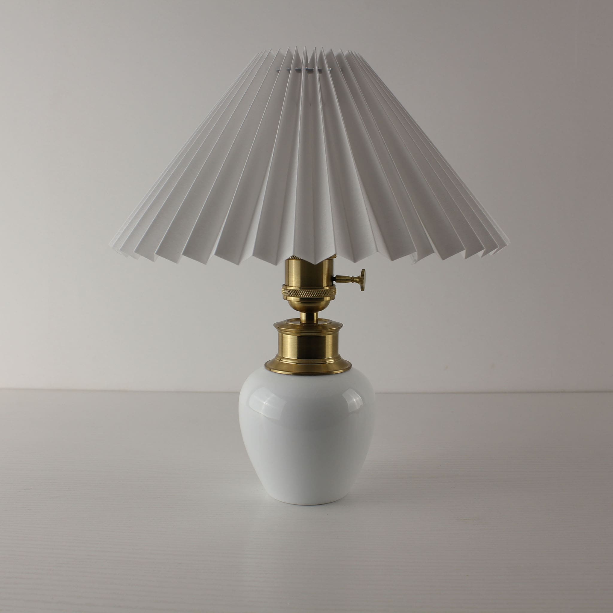 Minimalist white ceramic cordless lamp with a classic white pleated shade and brass toggle switch.