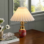Red ceramic cordless table lamp with pleated shade displayed on a wooden table beside a window