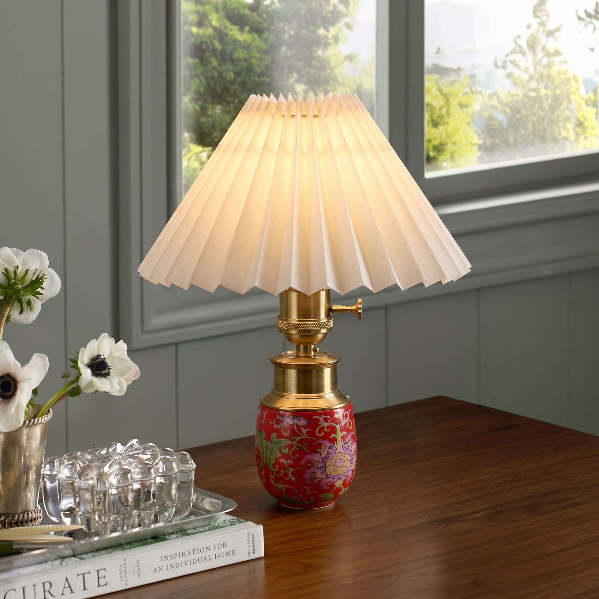Red ceramic cordless table lamp with pleated shade displayed on a wooden table beside a window
