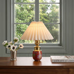 Red ceramic cordless table lamp with pleated shade displayed on a wooden table beside a window