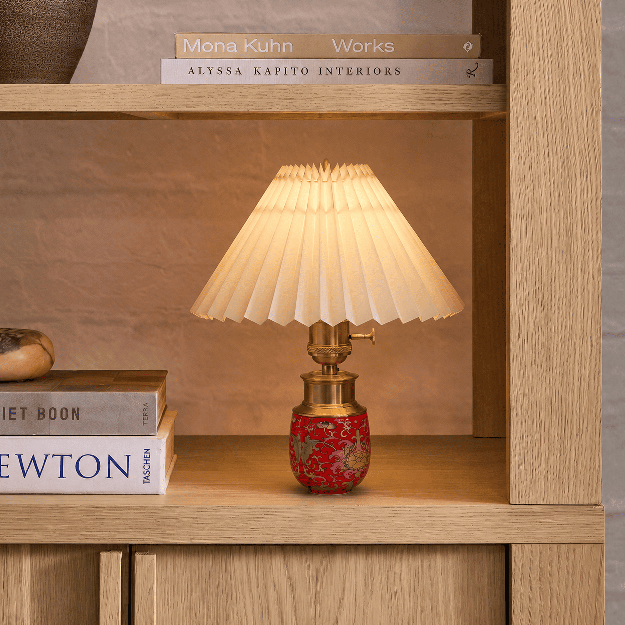 Red ceramic cordless table lamp with a pleated lampshade displayed on a wooden bookshelf