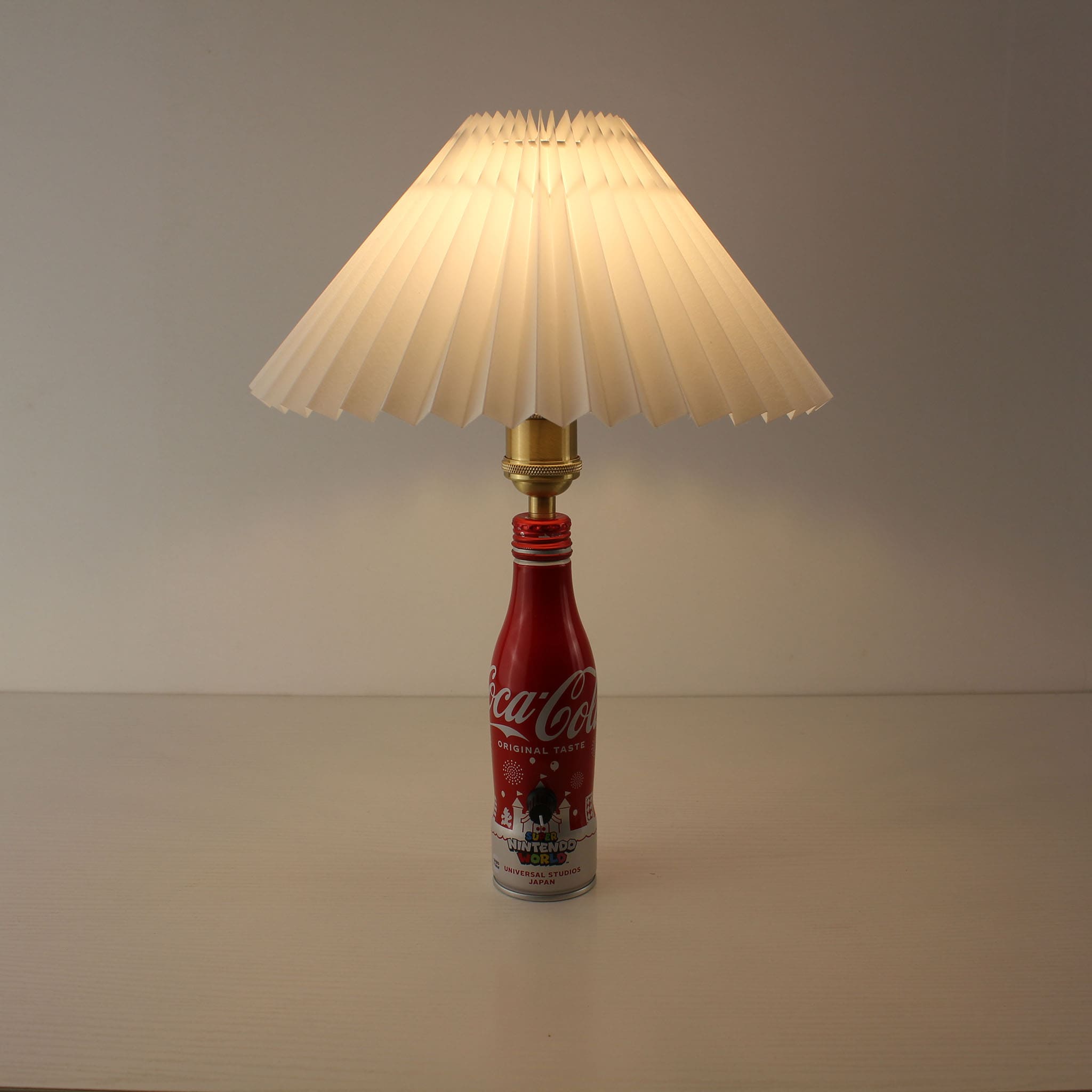 A cordless table lamp made from a red Super Nintendo World Coca-Cola bottle with a white pleated shade, shown in a bright studio setting